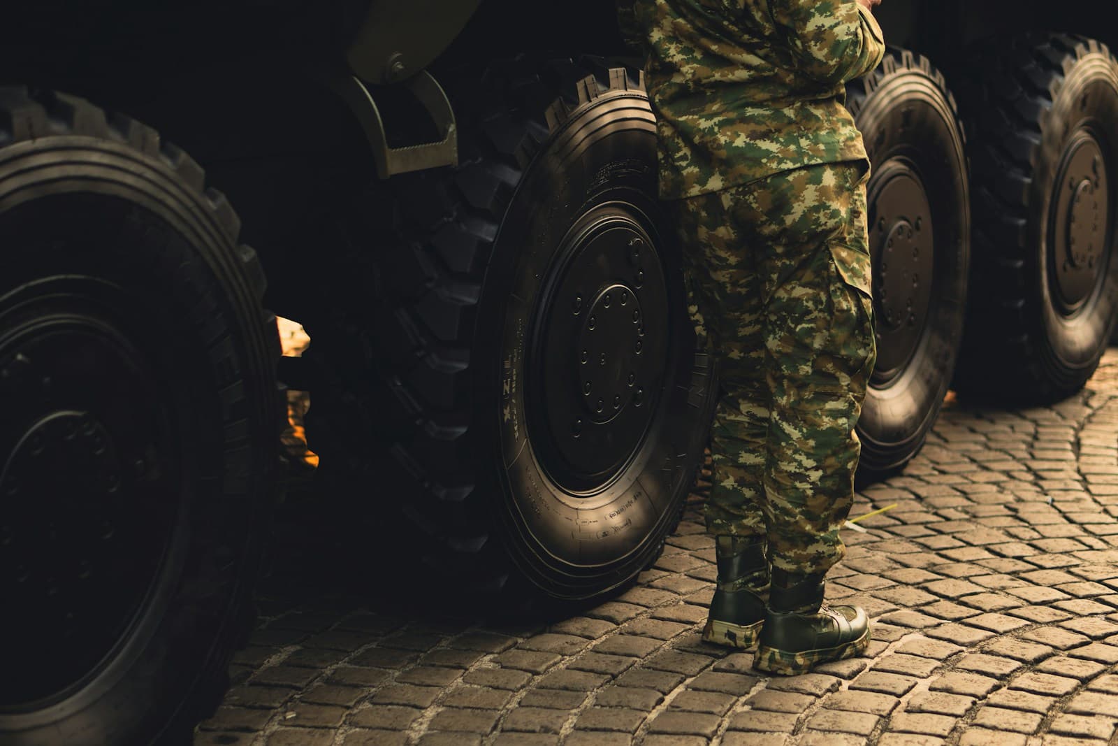 Service member in camouflage standing beside large military vehicle wheels